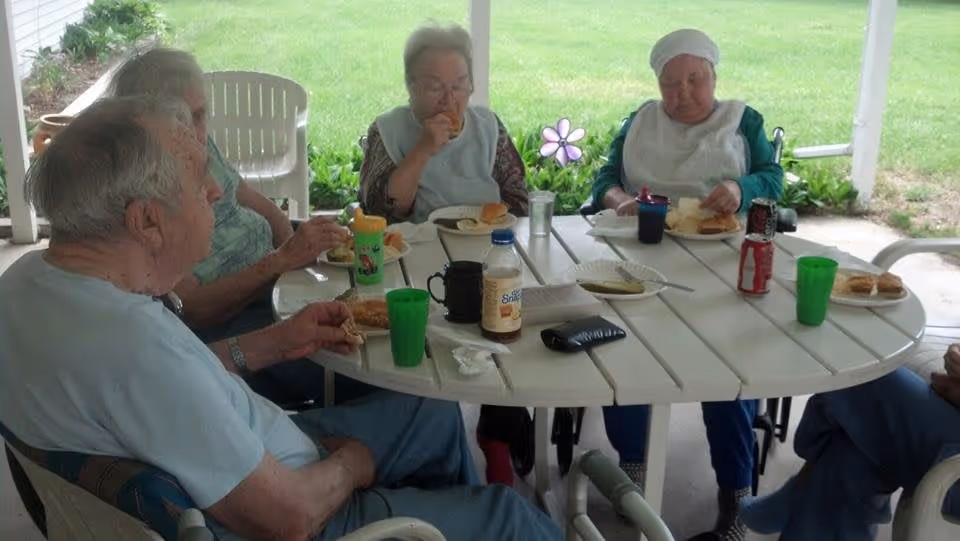 A group of elderly people sitting around a white outdoor table under a covered patio, eating a meal together. The table has plates with food, cups, and condiments. The background shows green grass and plants.