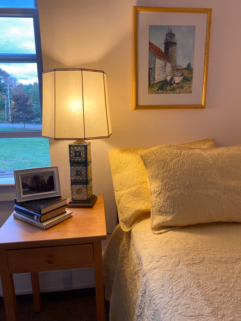 A cozy bedroom corner featuring a wooden nightstand with a decorative lamp, a stack of books, and a framed photo. Next to the nightstand is a bed with a textured yellow quilt and matching pillows. A window shows a green lawn and trees outside, and a framed painting of a lighthouse hangs on the wall above the bed.