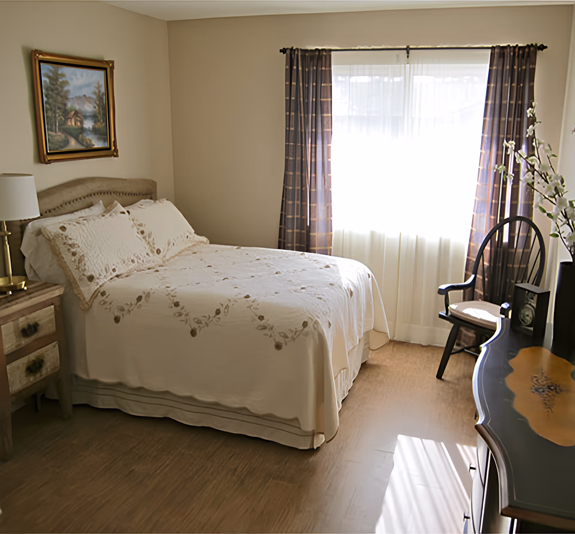 A cozy bedroom with a neatly made bed covered in a cream-colored quilt with floral embroidery. There is a wooden nightstand with a lamp on the left side of the bed, a framed painting of a cabin in the woods hanging on the wall above the bed, and a window with sheer white curtains and patterned drapes letting in natural light. A wooden chair and a decorative wooden dresser with a vase of white flowers are positioned near the window.