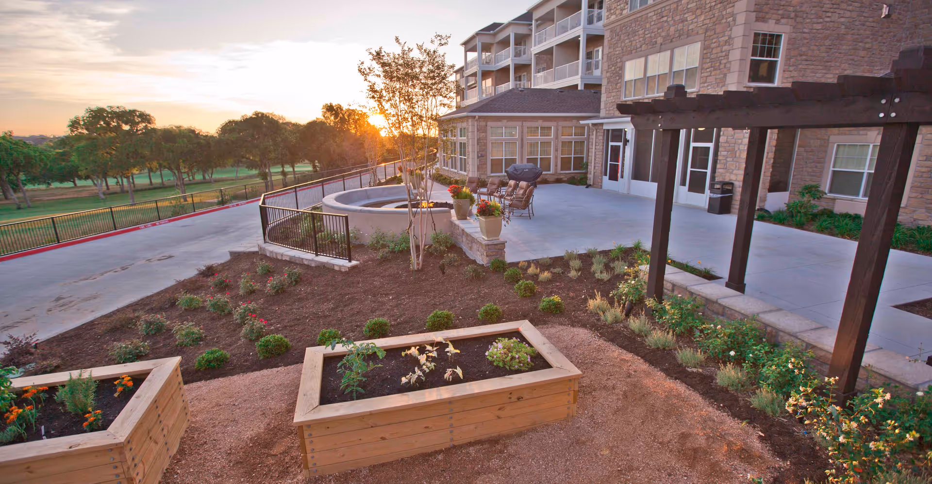Sunset view of a senior living facility's outdoor patio and landscaped garden with raised planter boxes, seating, and the building facade.