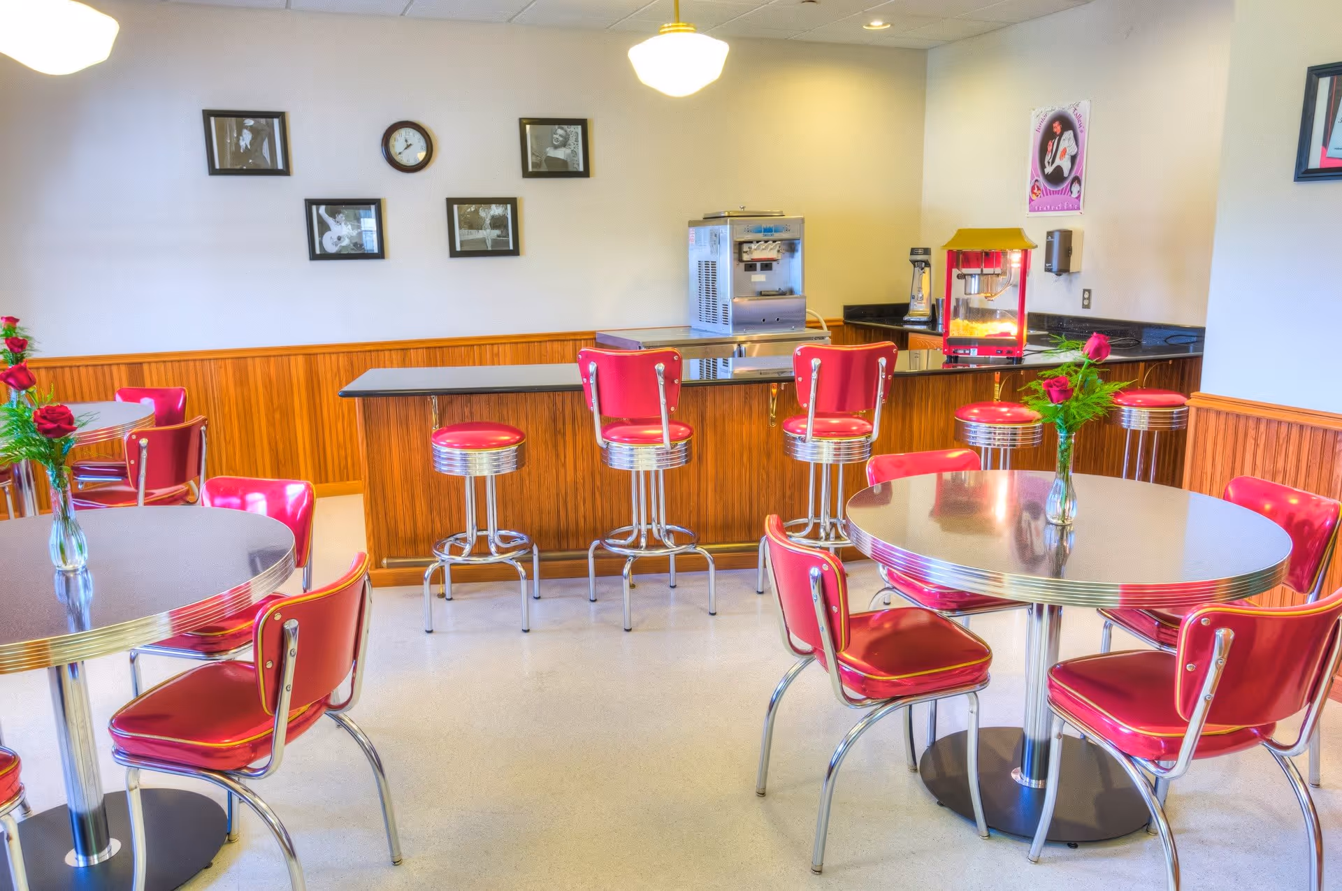 A retro-style dining area with round tables and red cushioned chairs. There is a counter with red bar stools, a popcorn machine, and a beverage dispenser. The walls have a few black and white framed pictures and a clock. Fresh flowers in vases are placed on the tables.