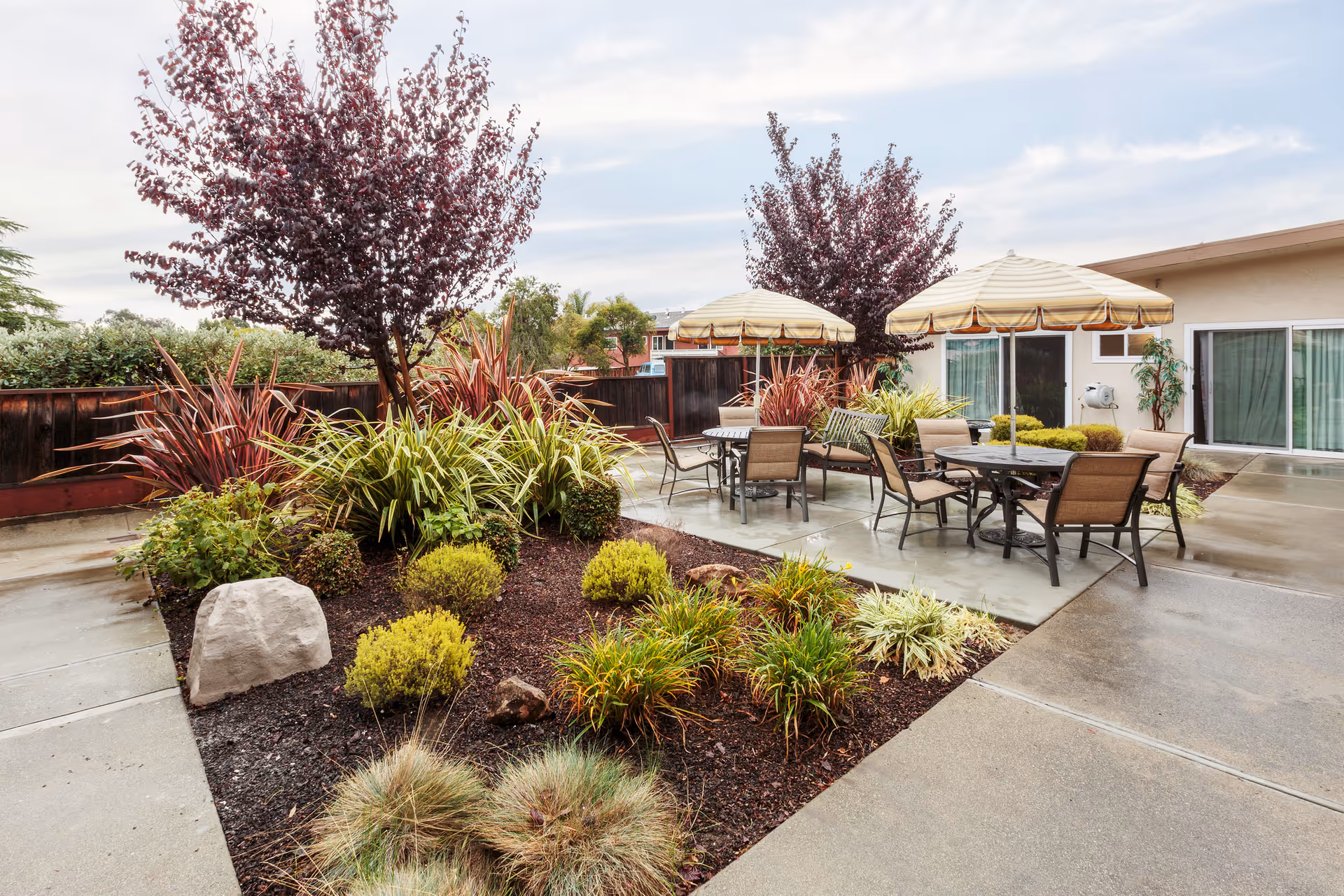 Outdoor patio area with tables and chairs under large beige umbrellas, surrounded by landscaped garden beds with various shrubs and small trees, adjacent to a building with sliding glass doors.