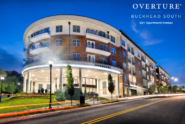 Exterior view of a modern multi-story apartment building at dusk with illuminated entrance and balconies. The building is labeled Overture Buckhead South, a 55+ apartment home community. The street in front has light trails from passing cars.