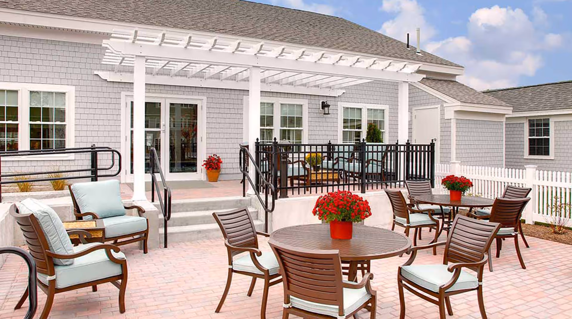 Outdoor patio area at Atria Woodbriar Place featuring multiple round wooden tables with cushioned chairs, red flower pots on the tables, a white pergola over a raised seating area with additional cushioned chairs, and a gray building with white trim in the background under a partly cloudy sky.