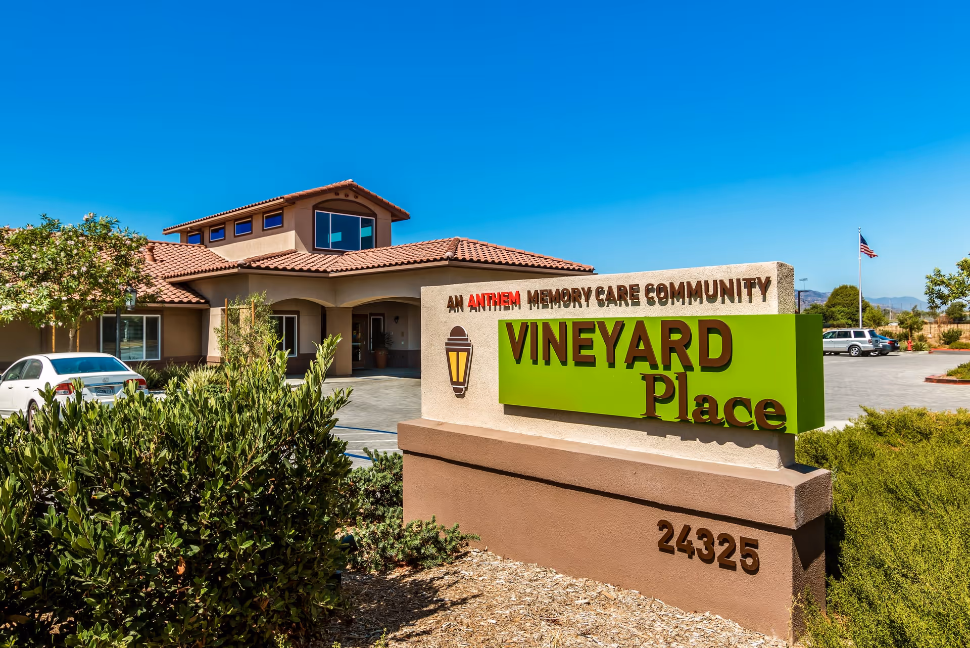 Exterior view of Vineyard Place, a memory care community building with a beige stucco facade and a red tile roof under a clear blue sky. In front, there is a large sign with green and brown lettering that reads 'Vineyard Place' along with the address number 24325. There are some bushes and trees around the entrance, a white car parked to the left, and an American flag flying in the background.