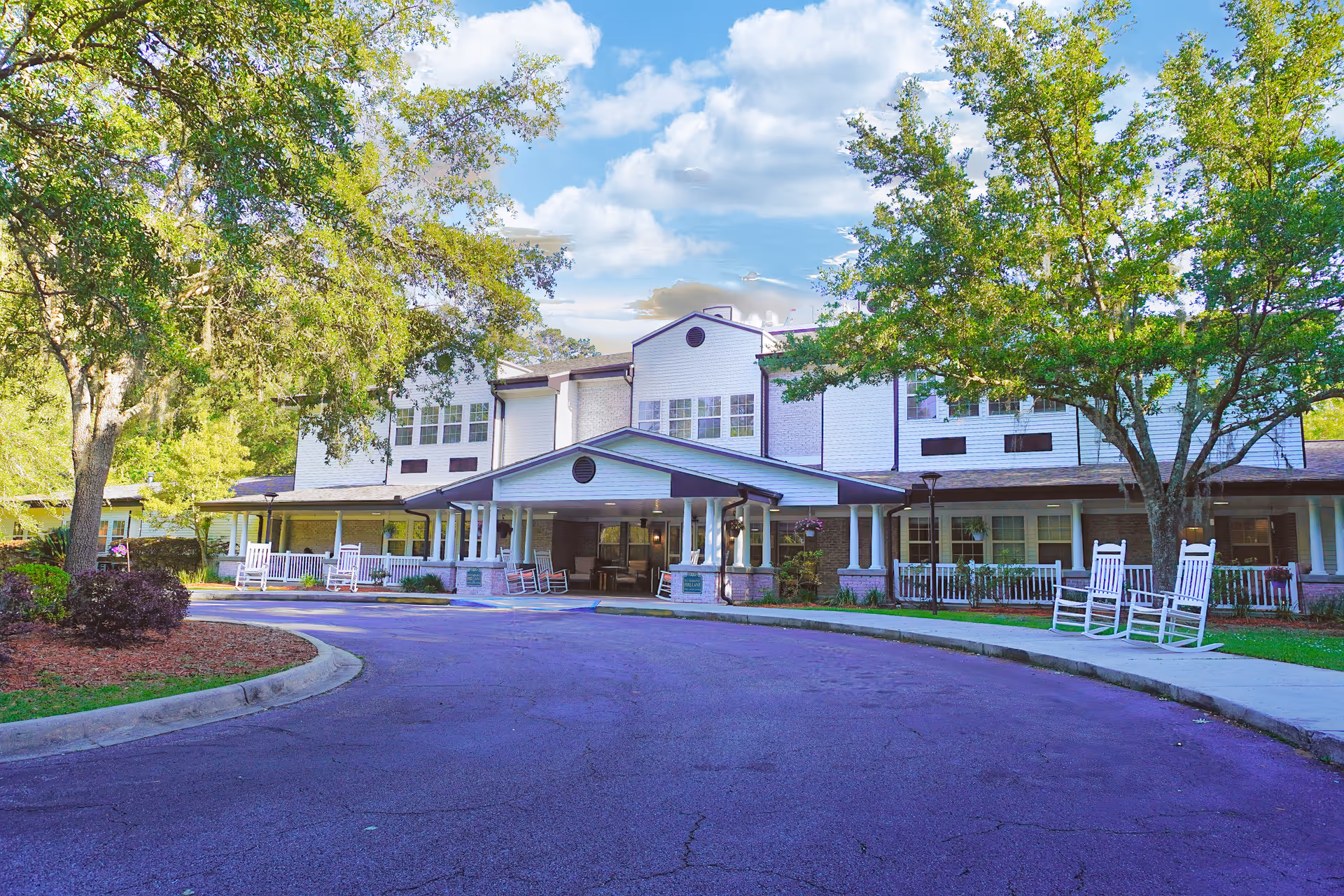 Front entrance of a two-story assisted living building with a covered porte-cochere, rocking chairs on the porch, and surrounding trees.