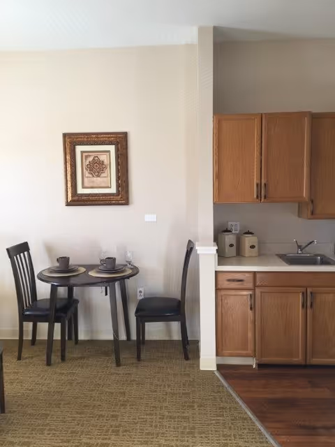 A small dining area with a round table set for two with placemats, cups, and glasses. Two black chairs are positioned around the table. Adjacent to the dining area is a kitchen with wooden cabinets, a countertop, a sink, and two canisters. A framed decorative artwork hangs on the wall above the dining table.