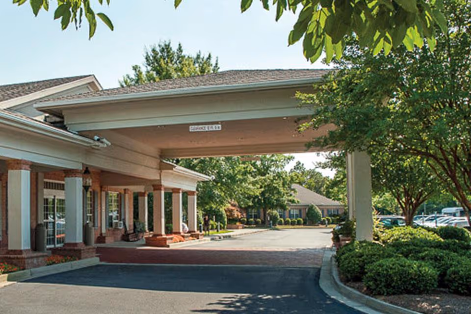 Entrance area of Life Care Center of Columbia, South Carolina showing a covered driveway with columns, surrounded by trees and shrubs, with a parking lot visible in the background.