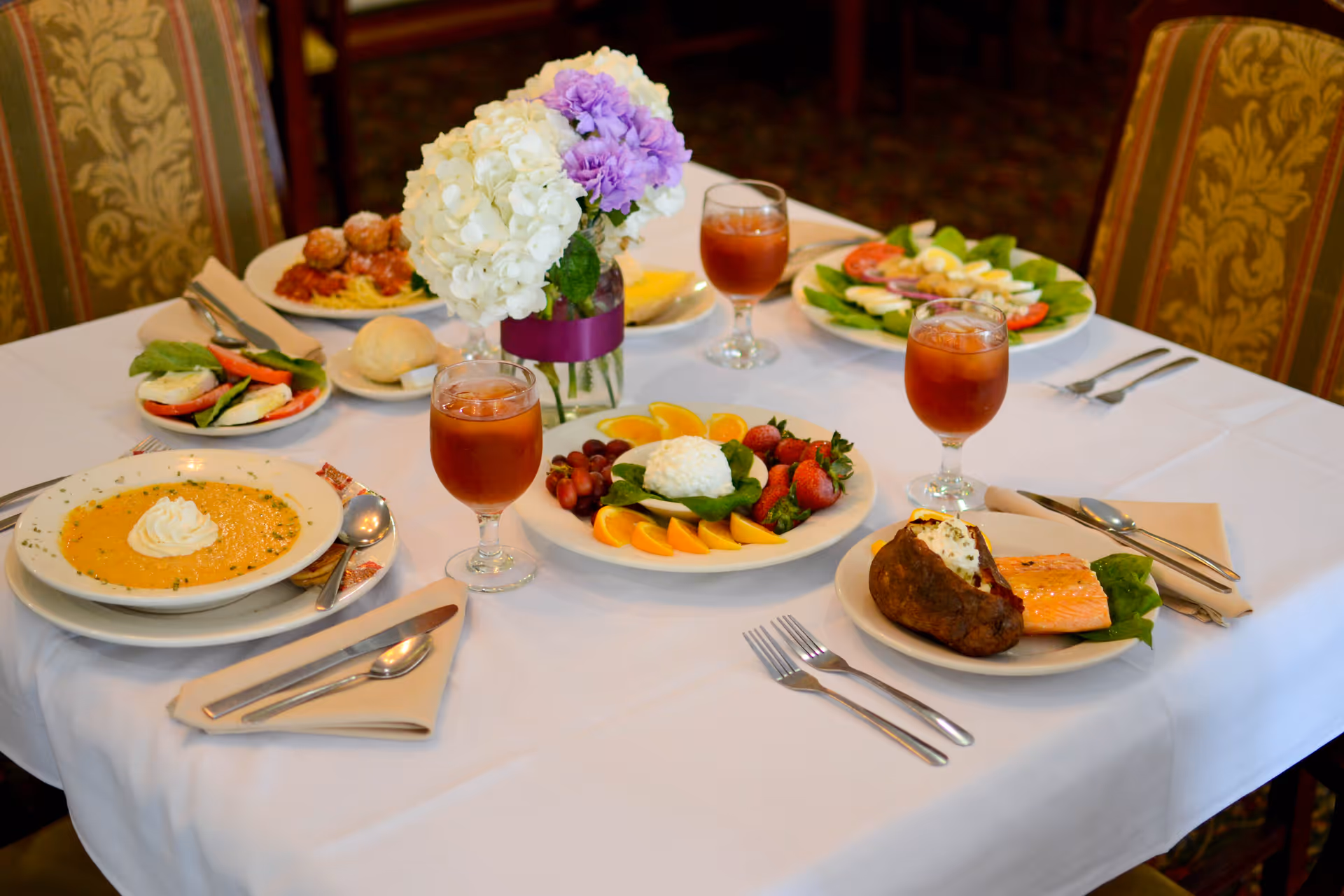 A dining table set with a white tablecloth and various plated meals including soup, salad, baked potato with salmon, fruit plate, and iced tea glasses. A vase with white and purple flowers is placed in the center of the table. The chairs around the table have patterned upholstery.