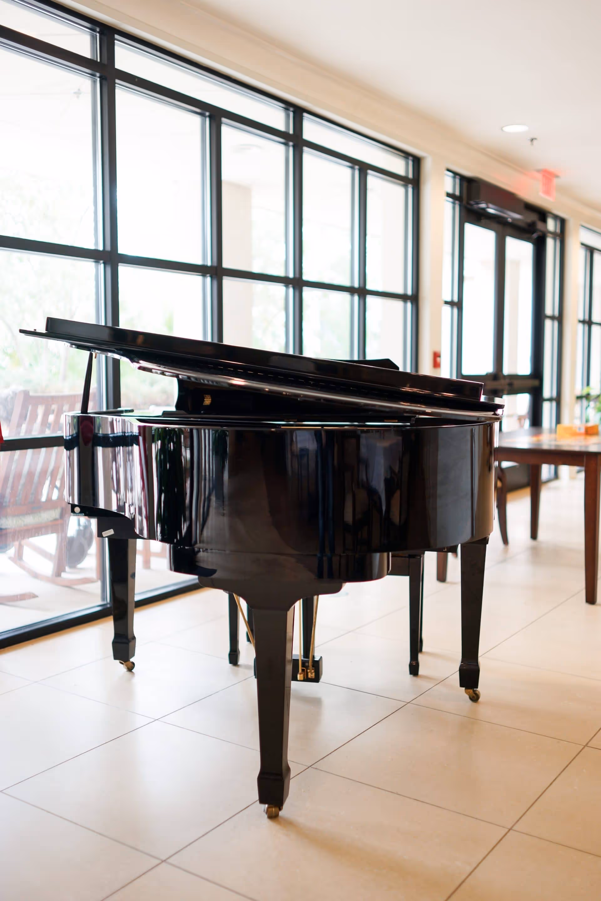 A shiny black grand piano with its lid open is placed in a bright room with large windows and tiled flooring. There is a wooden table and chairs visible in the background near the windows.