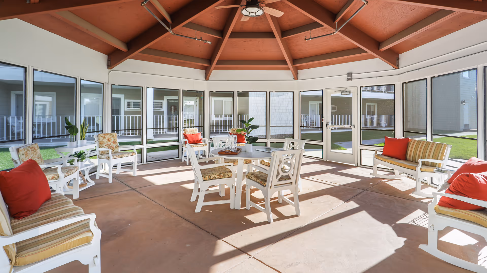 A bright, enclosed patio area with a high wooden ceiling and large windows surrounding the space. The room contains white wooden chairs and tables with floral and striped cushions, some with red accent pillows. There are potted plants on the tables and the floor, and the outdoor area with grass and building balconies is visible through the windows.
