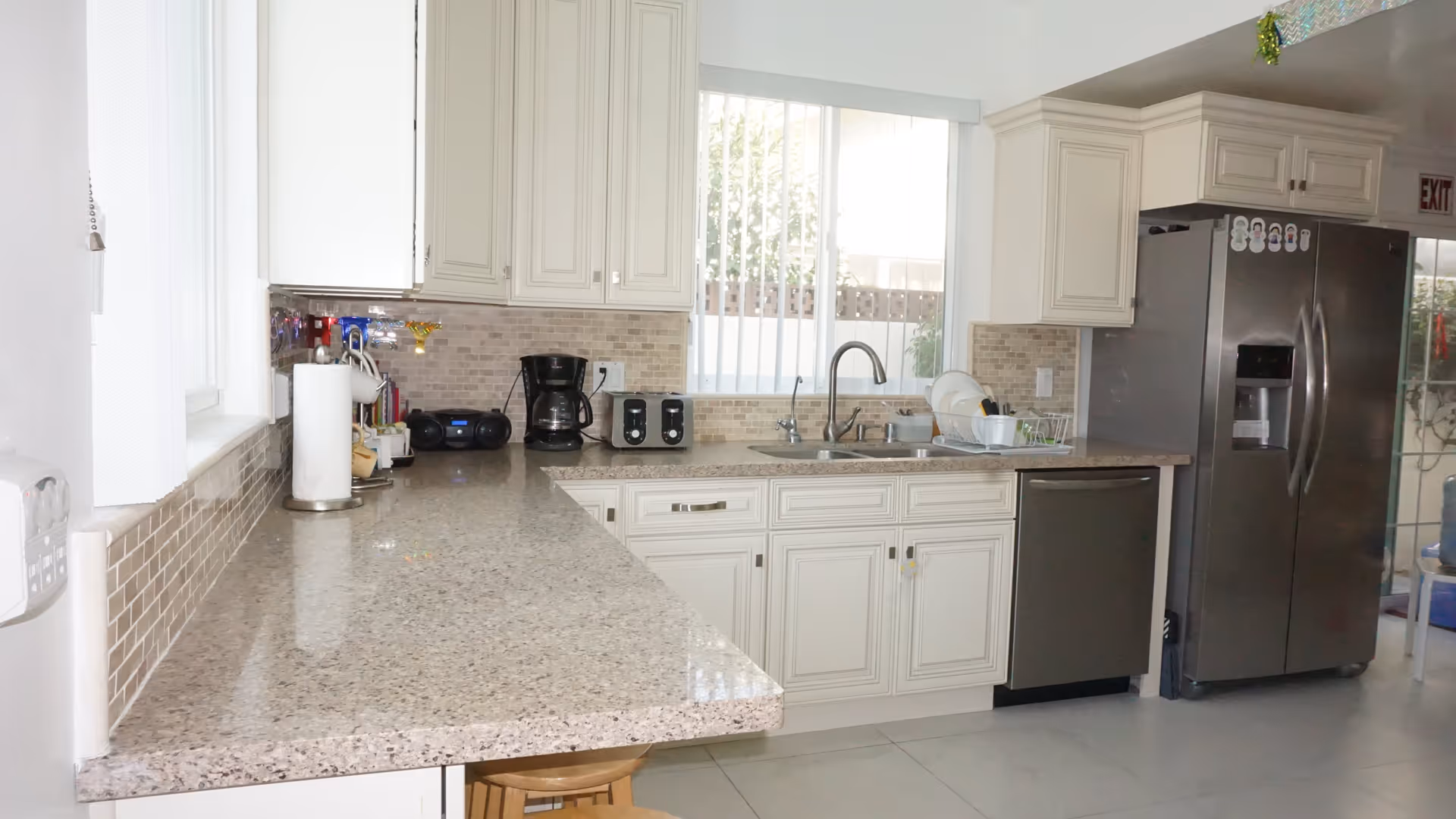 Bright and clean kitchen with beige cabinets, a large granite countertop, a double sink under a window with vertical blinds, a stainless steel refrigerator, dishwasher, coffee maker, toaster, and a paper towel holder.