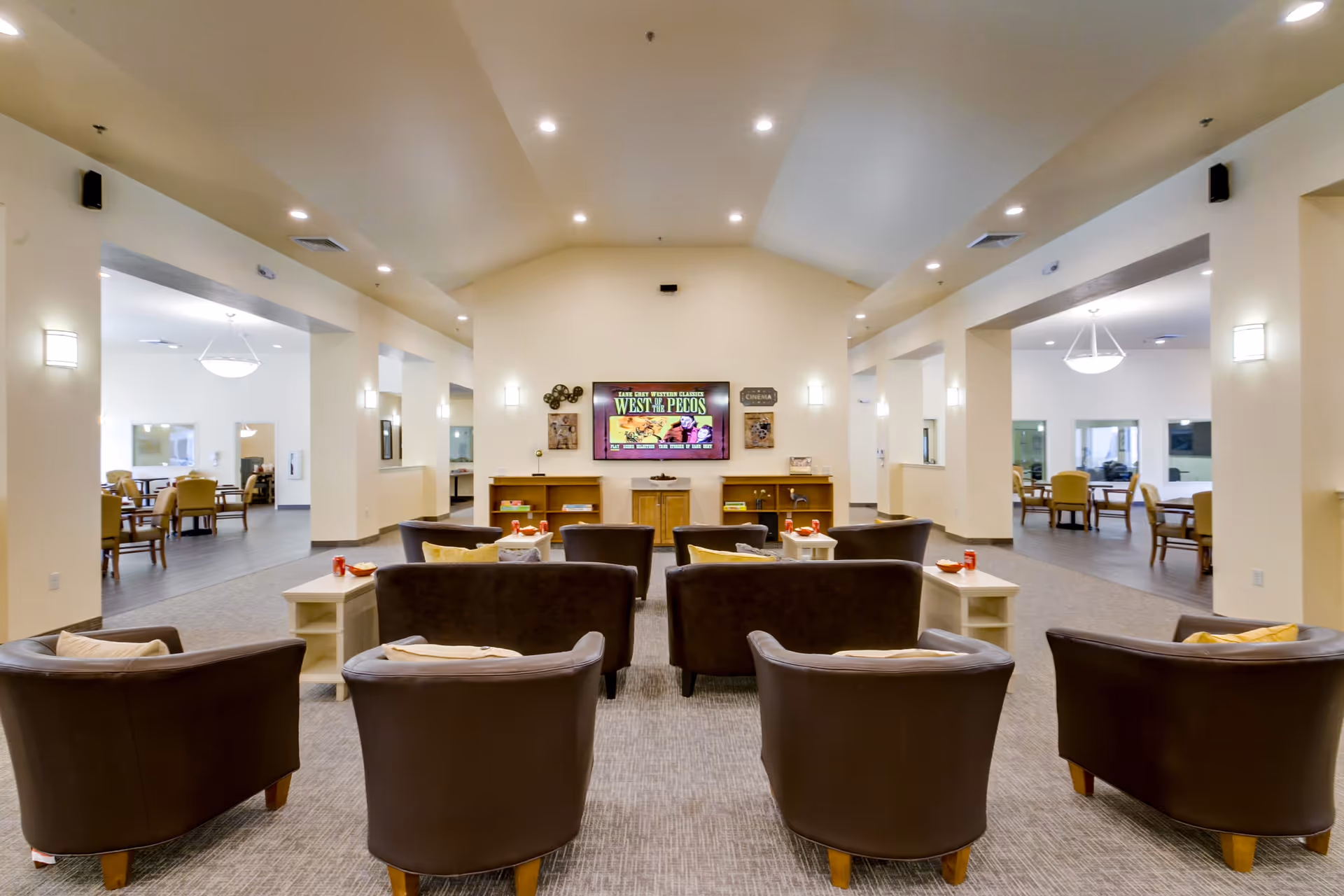 A spacious senior living common area with multiple brown armchairs and sofas arranged facing a large wall-mounted television displaying a western movie poster. The room has beige walls, recessed lighting, and side tables with snacks and drinks. Adjacent rooms with dining tables and chairs are visible through large open doorways on both sides.