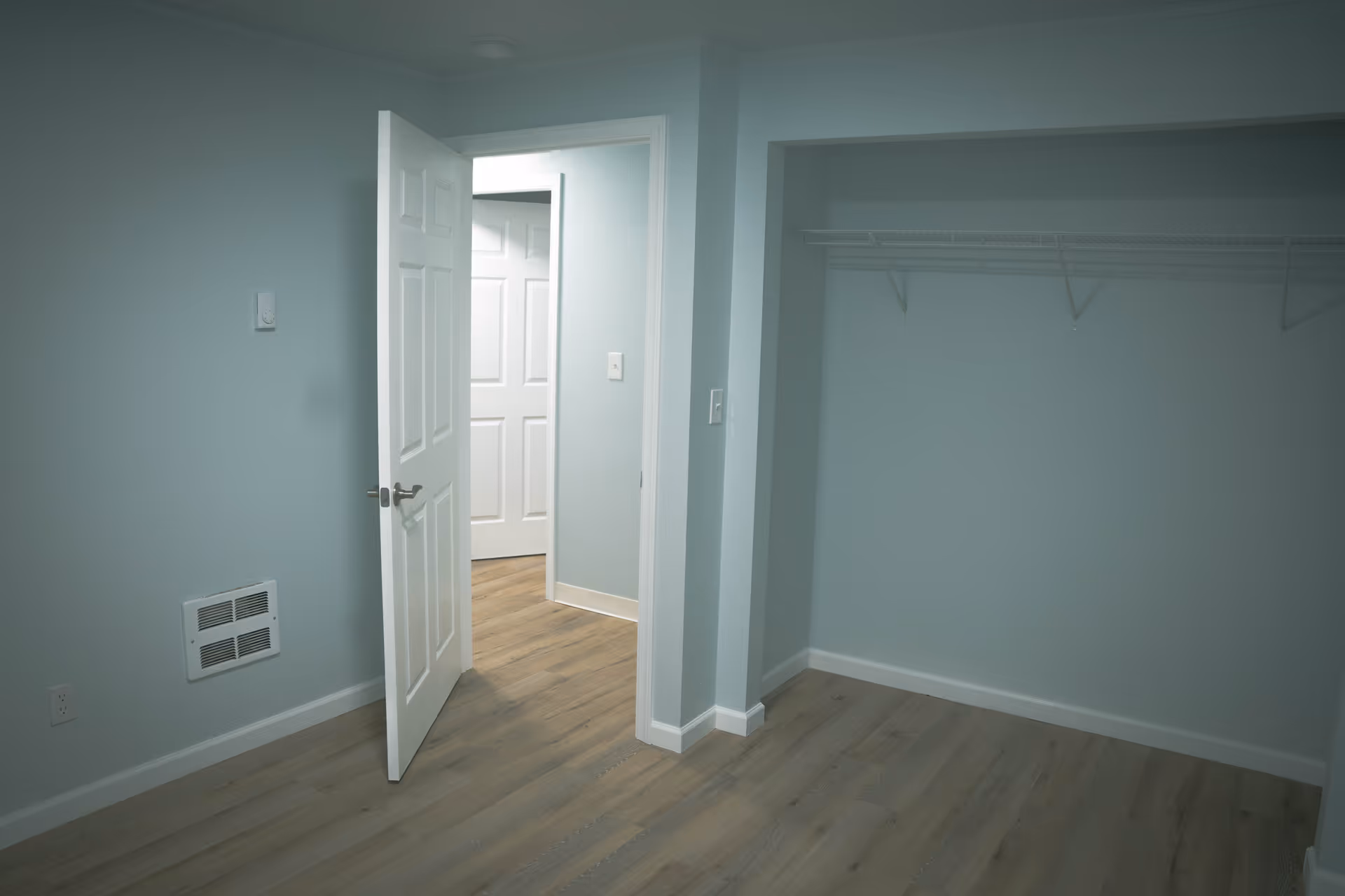 Empty room with light blue walls and wood flooring, featuring an open white door leading to a hallway with another white door. There is a large open closet space with a white wire shelf on the right side of the room.