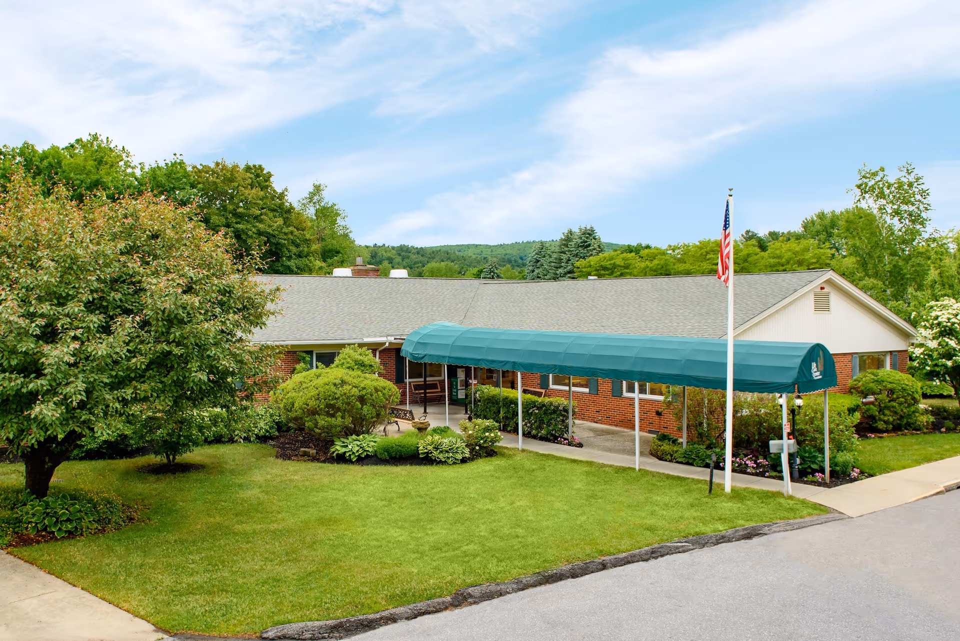 Front exterior of a single-story brick senior care building with a green awning-covered entrance, flagpole, and manicured lawn.