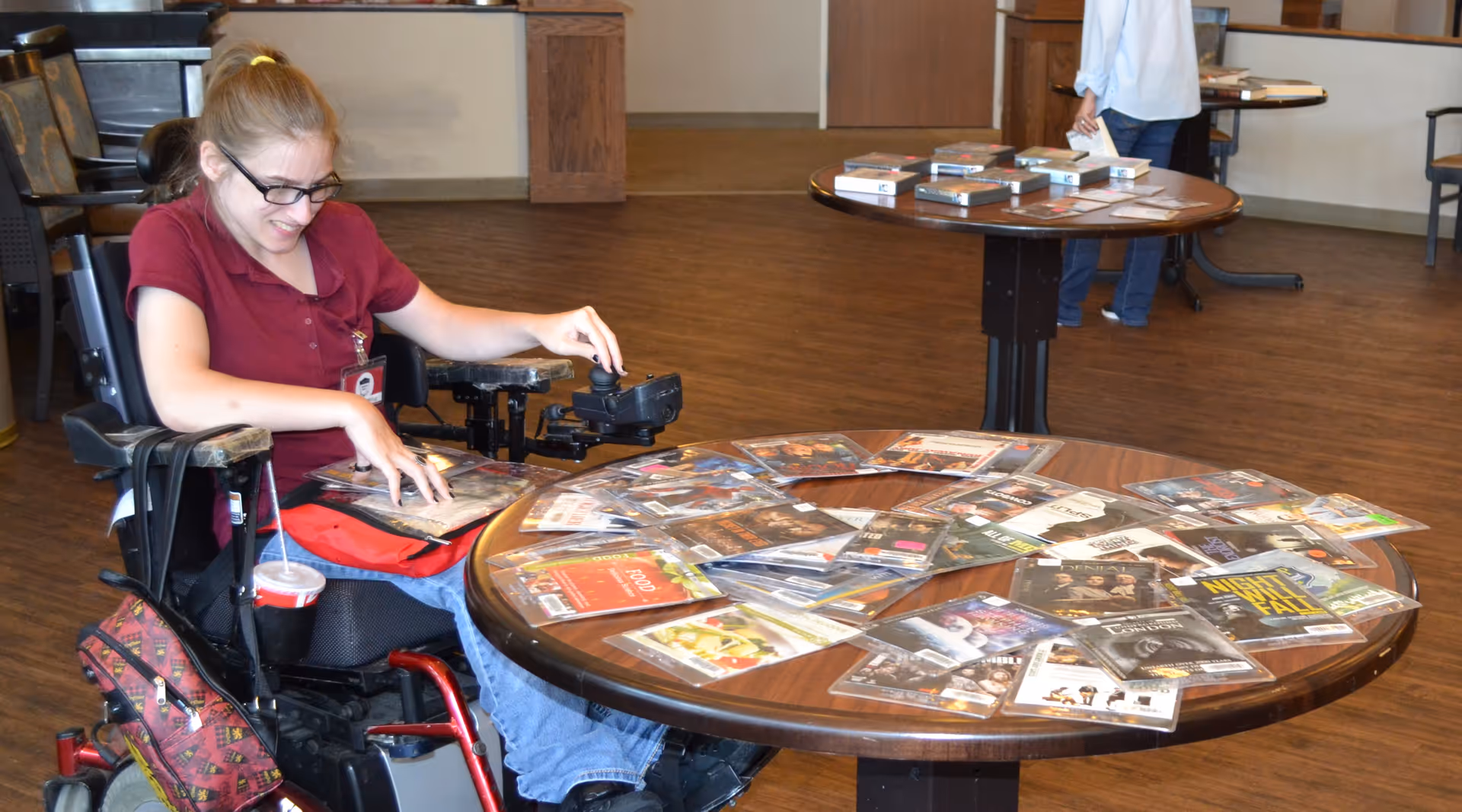 A woman in a wheelchair is selecting DVDs from a round wooden table covered with various DVD cases in a spacious room with wooden flooring. Another person is visible in the background near a second table with more DVDs.