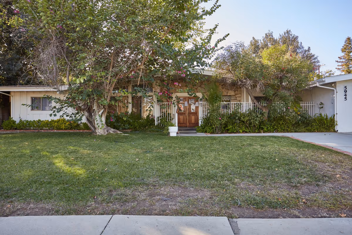 Front exterior view of a single-story building with a wooden double door entrance, surrounded by lush greenery and trees. There is a well-maintained lawn in front and a concrete driveway to the right. The building number 5945 is visible on the right side.