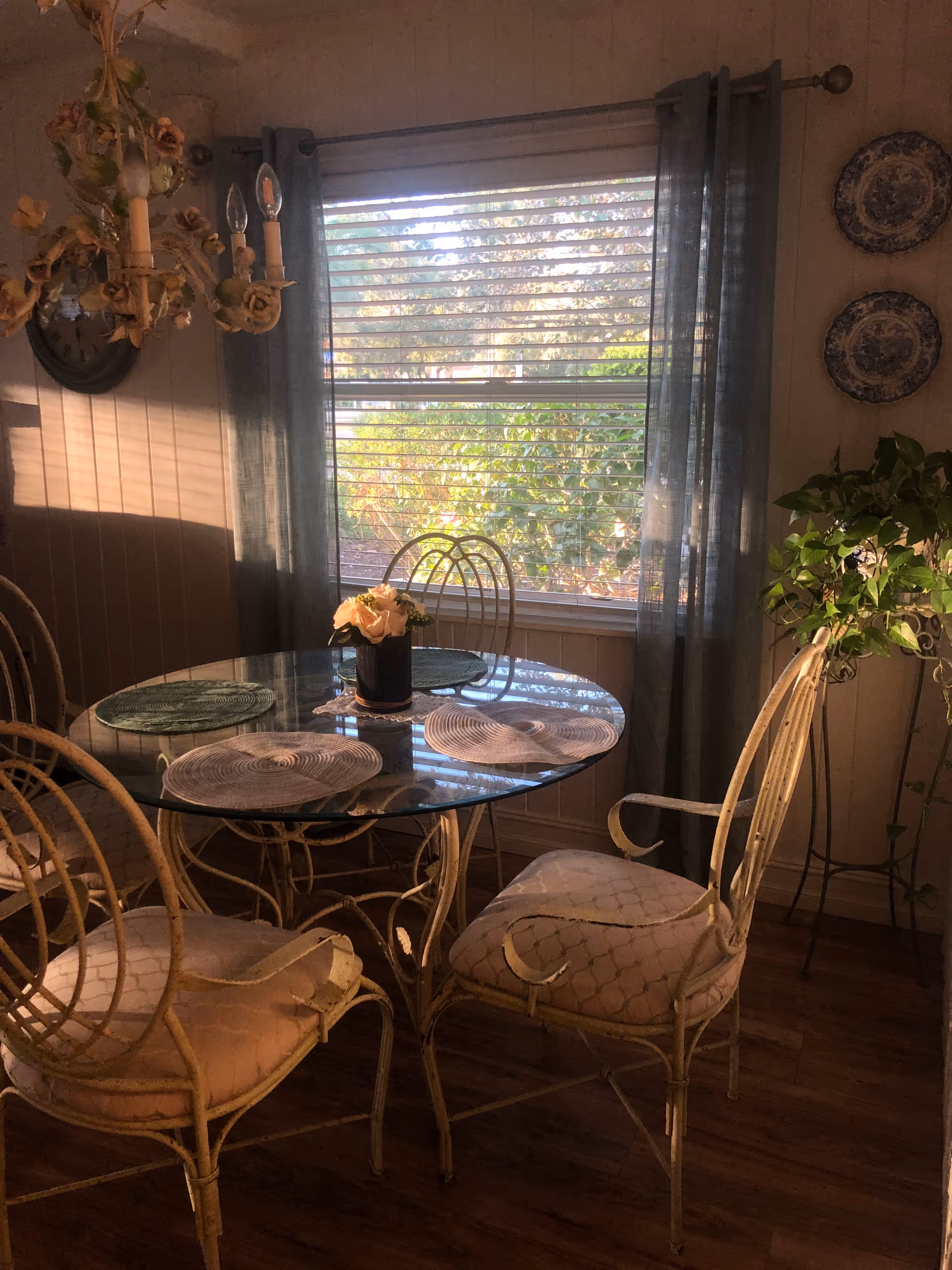 Sunlit dining area with a round glass-top table surrounded by four wicker chairs, a window with blinds and curtains, and a potted plant.