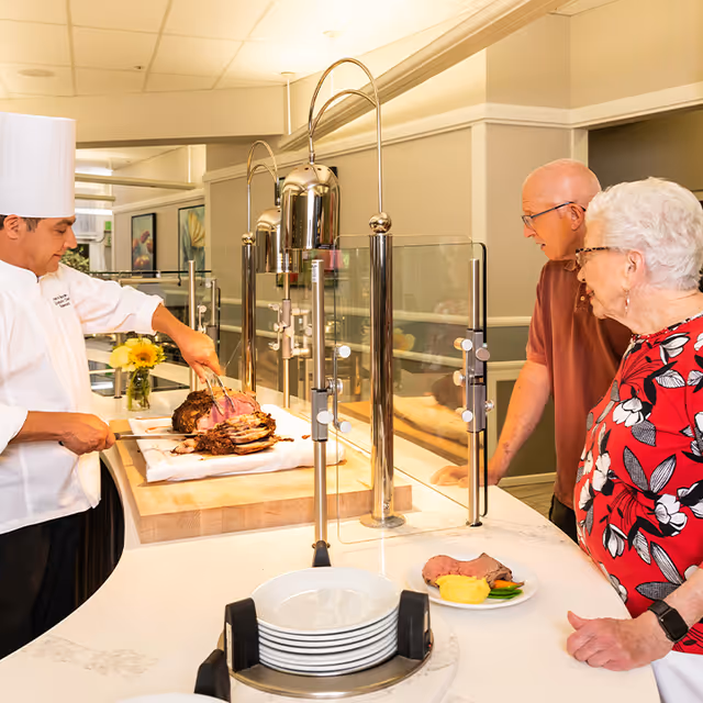 A chef carves a roast behind a service counter while two elderly residents watch and receive plated food in a dining area.