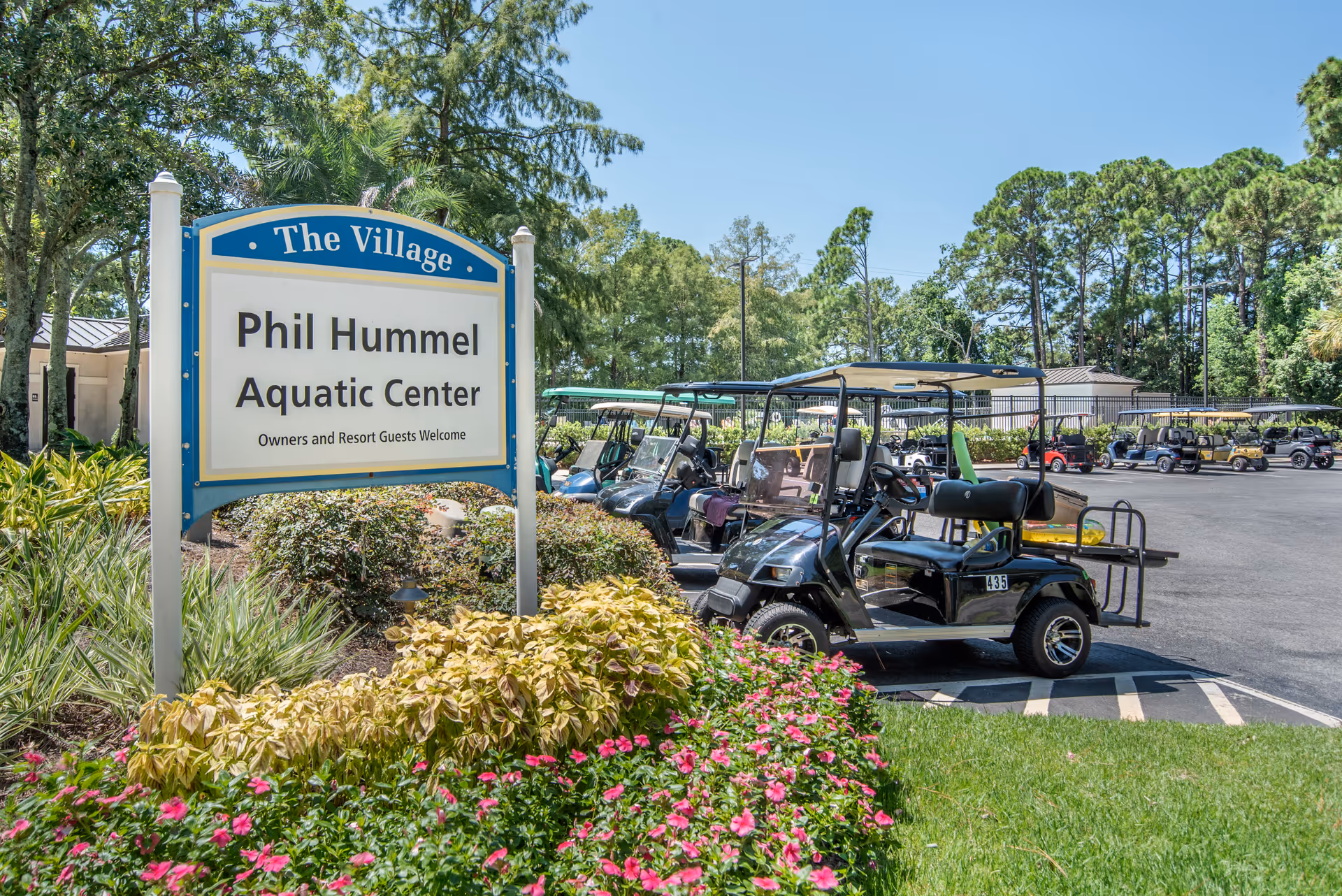 Sign reading 'The Village Phil Hummel Aquatic Center' beside landscaped flower beds and several parked golf carts in a sunny outdoor area.