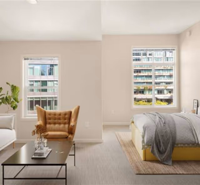 A bright and modern room with a beige armchair, a white sofa, and a black coffee table with decorative items. The room has two large windows showing an urban building outside. On the right side, there is a bed with gray bedding and a beige rug underneath.