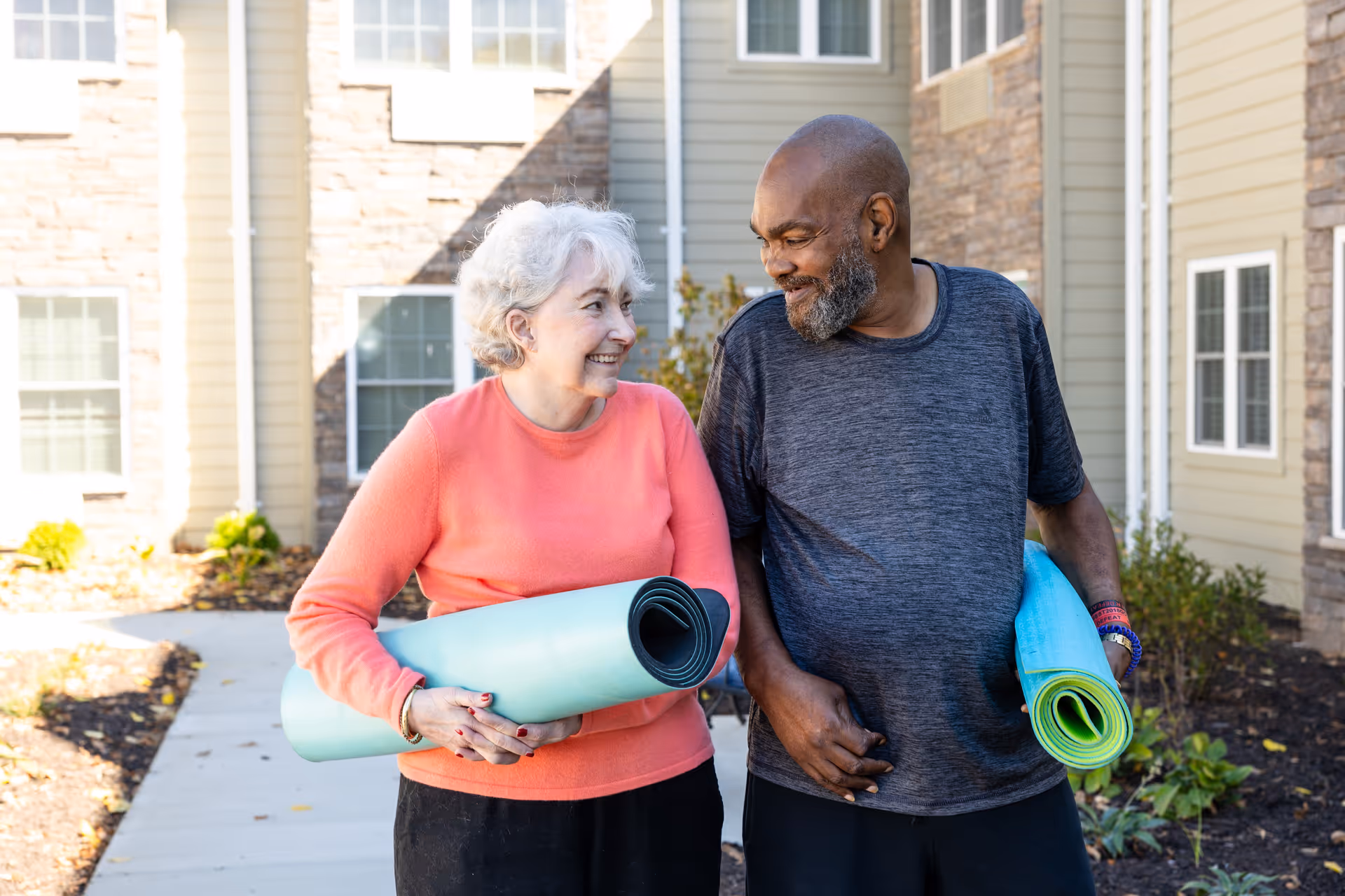 An elderly woman and man smiling at each other while holding rolled-up yoga mats, standing outside a senior living facility with beige siding and stone accents.
