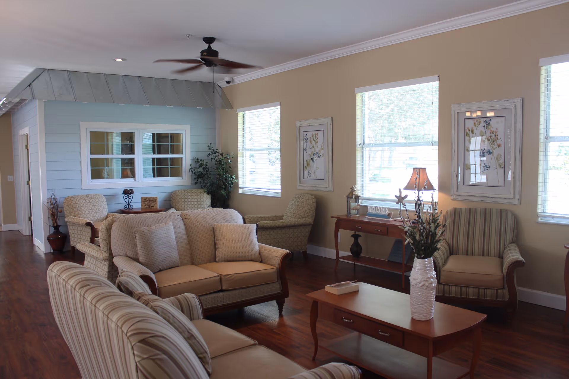 A cozy living room area in an assisted living facility featuring multiple upholstered armchairs and sofas arranged around a wooden coffee table with a white vase of flowers. The room has beige walls, three large windows with blinds, framed floral artwork, a wooden side table with a lamp, and a ceiling fan. The floor is wooden, and there is a small blue accent wall with a window and two additional chairs in the background.