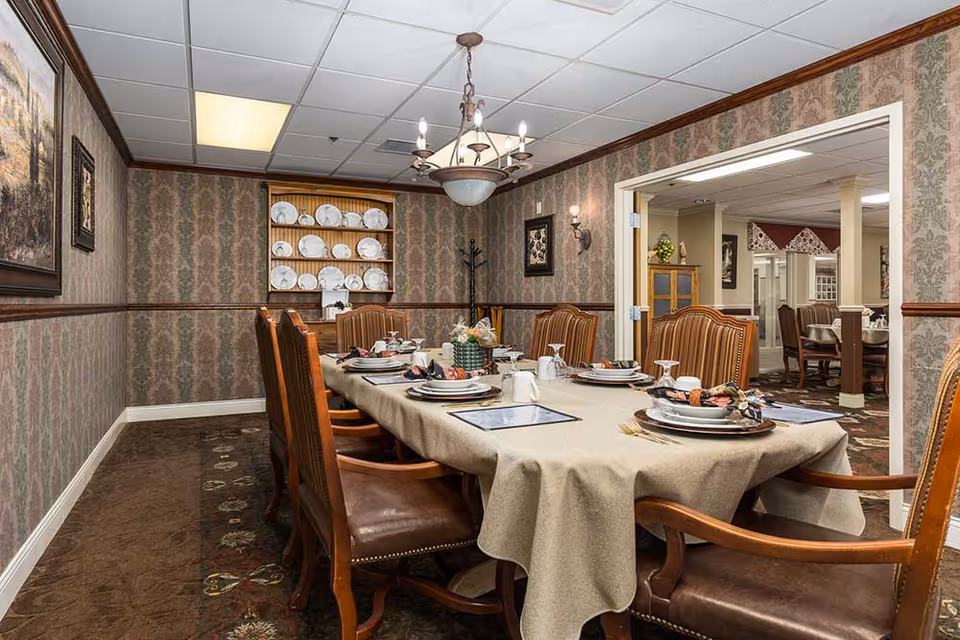 A formal dining room with a long table covered by a beige tablecloth, set with plates, napkins, glasses, and silverware. The room has patterned wallpaper, a chandelier hanging from the ceiling, and a cabinet displaying white plates. There are wooden chairs with leather seats around the table, and an open doorway leads to another dining area with similar decor.
