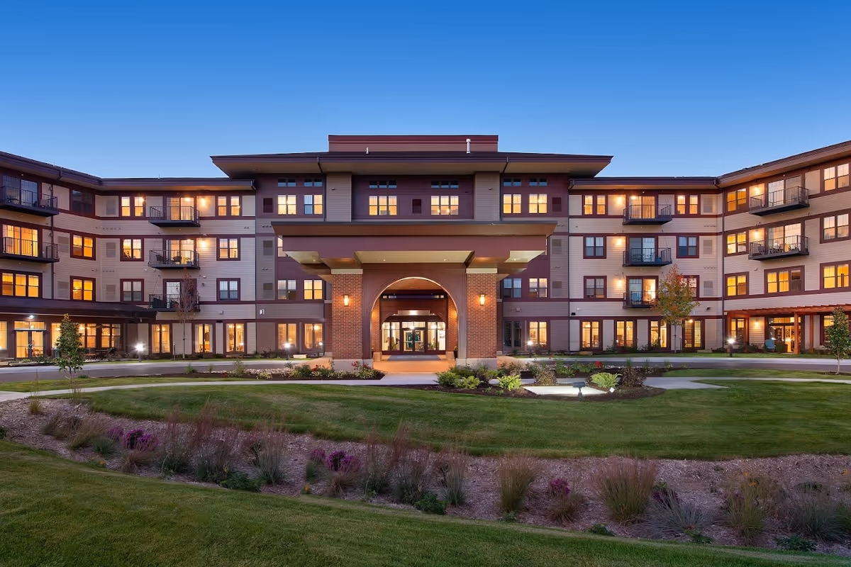 Exterior view of a large, modern senior living facility building at dusk with lights glowing from the windows. The building has multiple floors with balconies and a covered entrance. Well-maintained landscaping with grass, shrubs, and small trees is visible in the foreground under a clear blue sky.