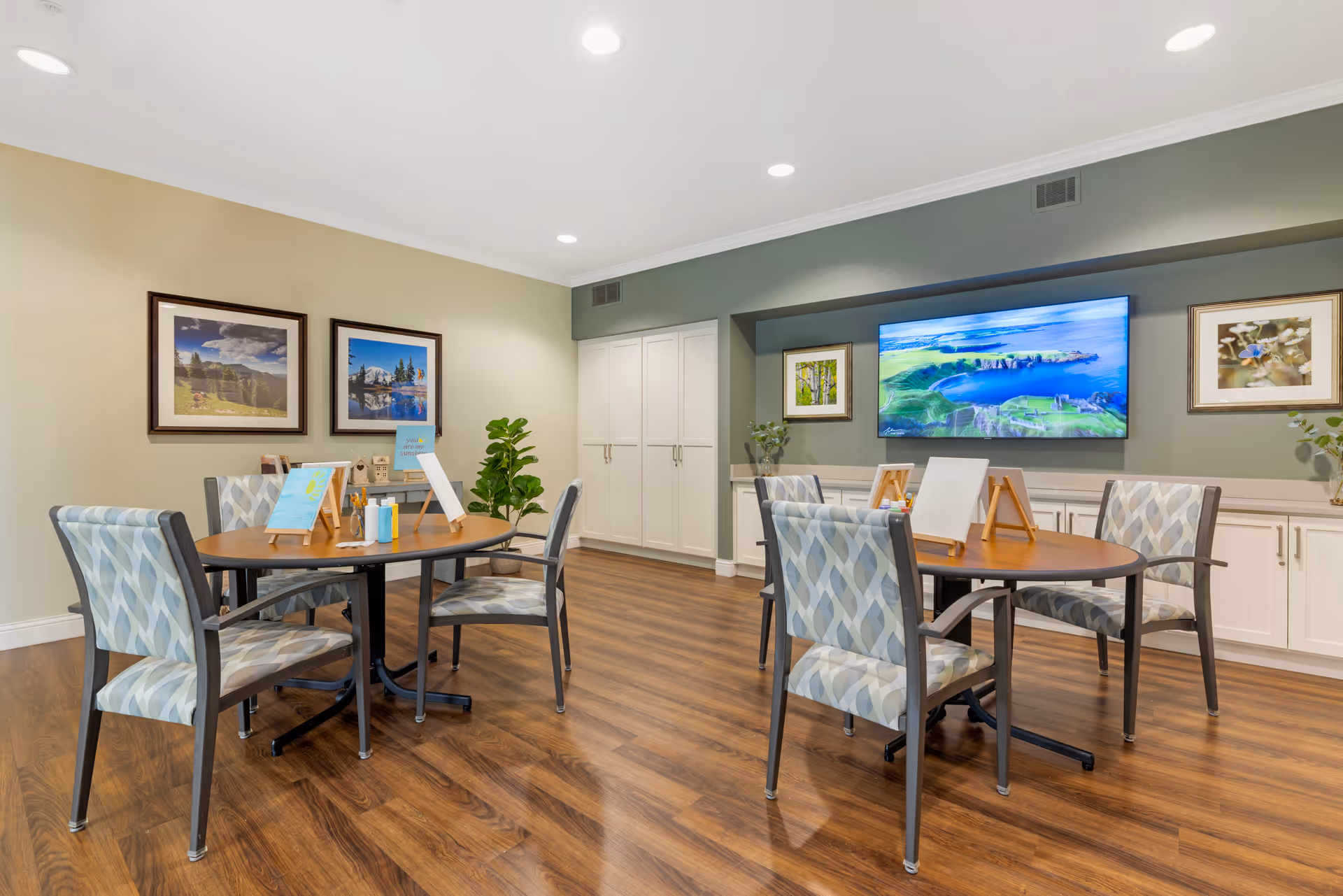 A bright and clean activity room with two round tables surrounded by patterned chairs. Each table has small easels with blank canvases and painting supplies. The room has wood flooring, light-colored walls, framed nature photographs, a large flat-screen TV mounted on a green accent wall, and white cabinetry along the back wall.