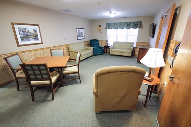A cozy living room area with a beige armchair in the foreground, two light green sofas, a dark green recliner, a wooden table with four chairs, a side table with a lamp, framed pictures on the walls, and a window with green valance curtains letting in natural light.