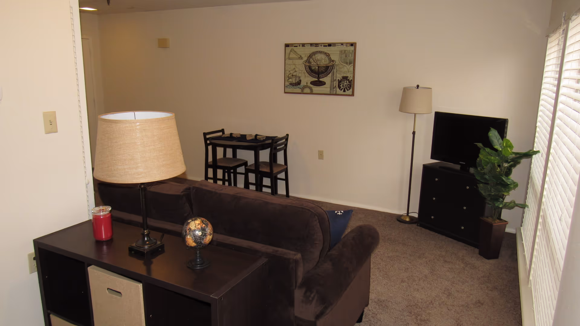 A living room area with a dark brown sofa facing a TV on a black dresser. There is a small black table with a lamp, a red candle, and a decorative globe behind the sofa. In the background, there is a small dining table with two chairs and a framed picture on the wall. A floor lamp and a potted plant are next to the TV. The room has beige carpet and white walls with a window covered by blinds on the right side.