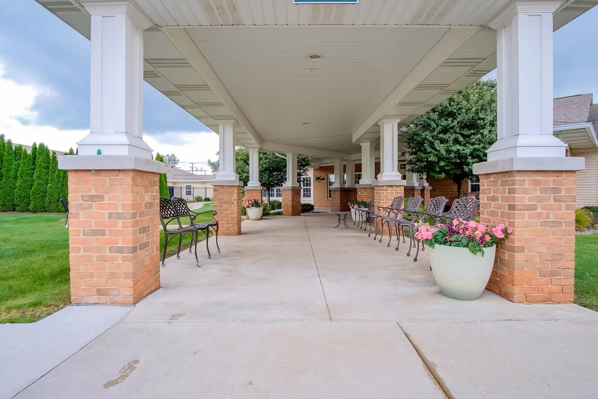 Covered outdoor walkway with brick pillars and white columns, lined with metal benches and large flower pots containing pink flowers, adjacent to a green lawn and trees under a cloudy sky.