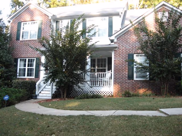 Front exterior view of a two-story brick house with white trim and green shutters. There is a small porch with white railings and a tree partially obscuring the entrance. A curved concrete walkway leads to the porch, and there is a grassy lawn in front.