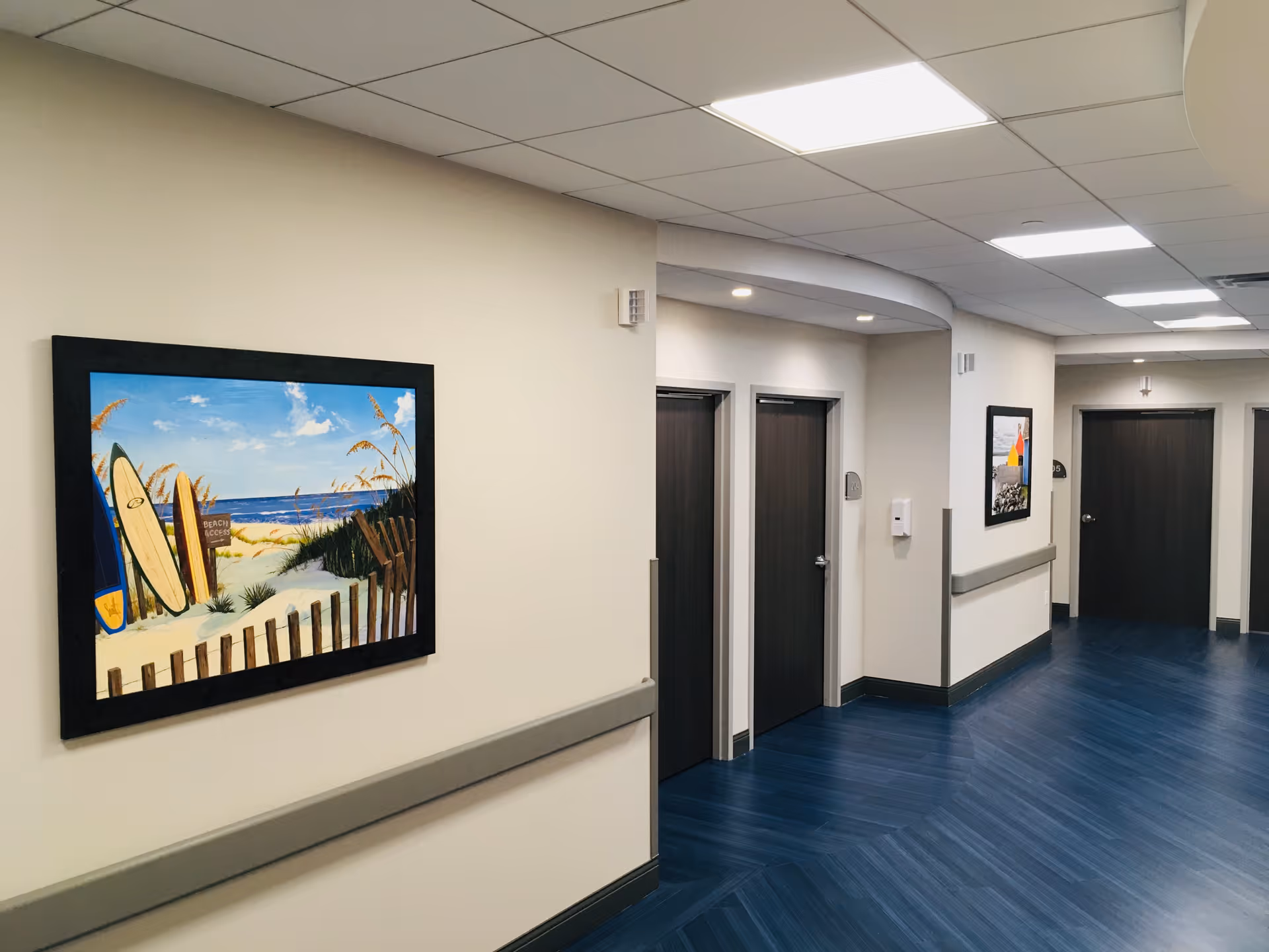 A clean, well-lit hallway in a nursing and rehabilitation center with blue flooring and beige walls. The hallway features several closed dark wooden doors and two framed paintings on the walls, one depicting a beach scene with surfboards and the other a lighthouse. There are handrails along the walls and ceiling lights illuminating the corridor.