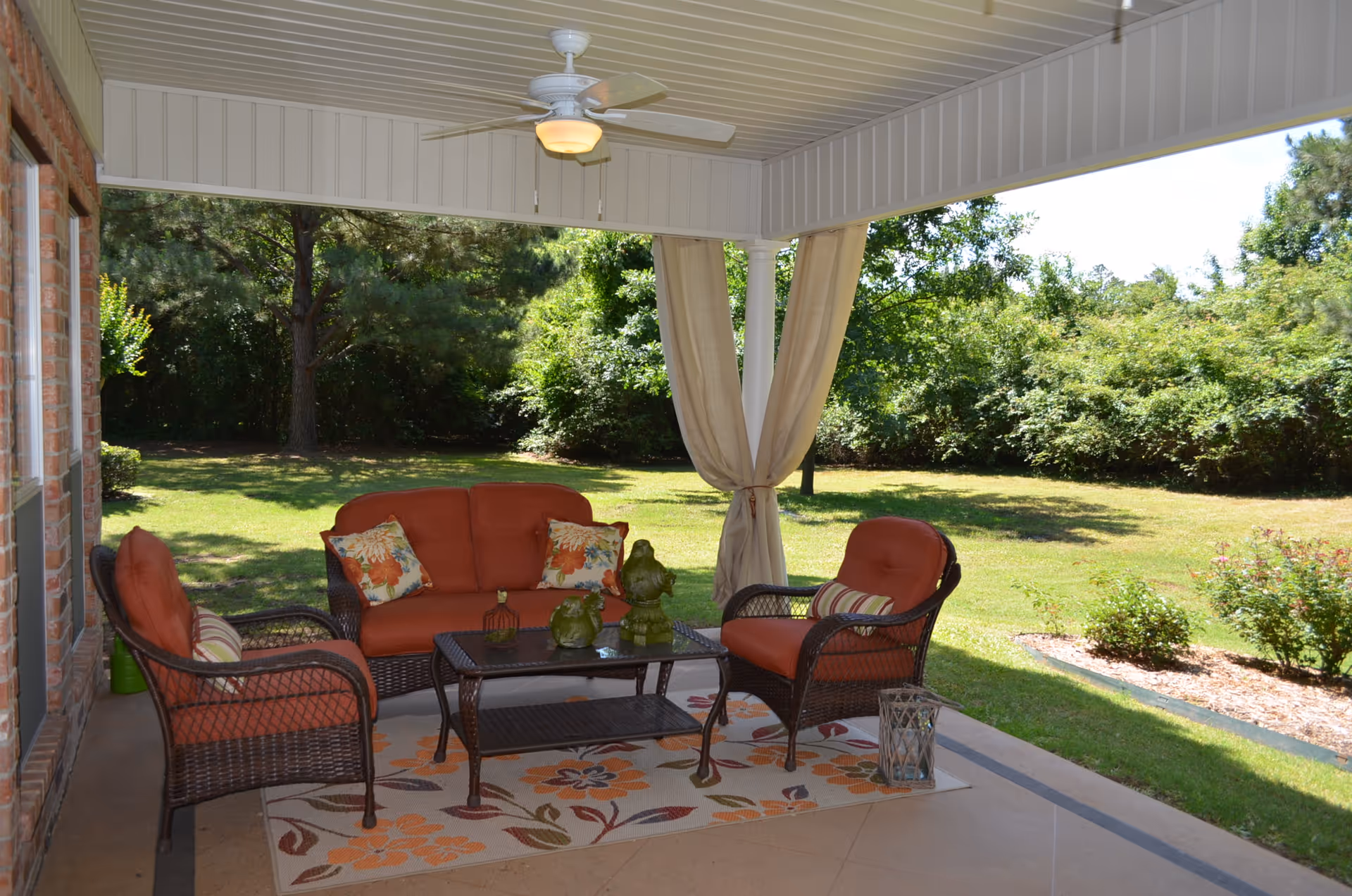Covered outdoor patio area with wicker furniture including a loveseat and two chairs with orange cushions and floral pillows. A glass-top coffee table sits on a floral patterned rug. The patio overlooks a grassy yard with trees and shrubs, and there is a ceiling fan above.