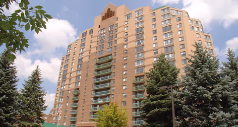Multi-story brick residential building with balconies surrounded by evergreen trees under a partly cloudy sky.
