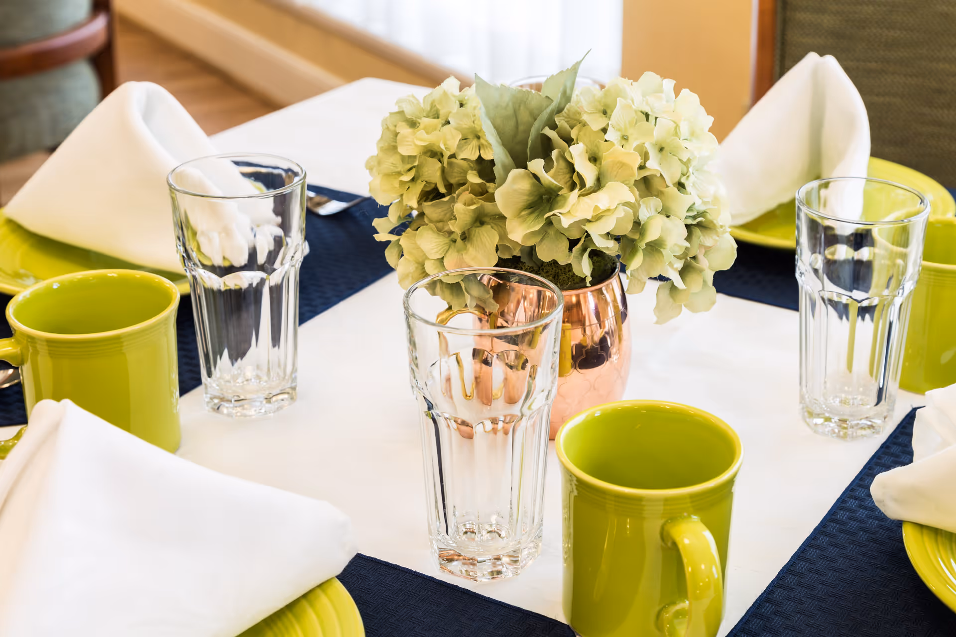 A close-up of a dining table set with green mugs, clear drinking glasses, white folded napkins on green plates, and a centerpiece of light green hydrangea flowers in a copper vase.