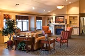 A cozy living room area in a senior living facility featuring comfortable seating including armchairs and a sofa arranged around a coffee table. The room has a fireplace with a painting above it, built-in shelves with decorative items, large windows with valances, and warm lighting from ceiling fixtures. There is a plant near the window and a patterned carpet covering the floor.