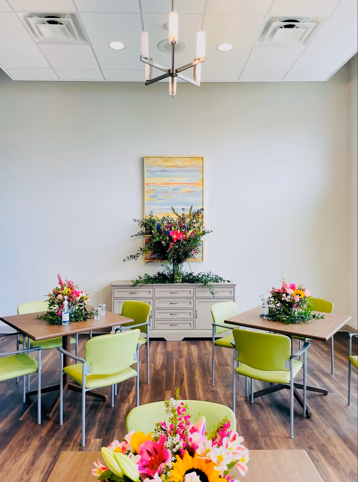 A bright dining room with light green chairs around wooden tables, each table decorated with colorful floral arrangements. A sideboard with a large flower arrangement and a framed painting is against the back wall. The room has wood flooring and a modern ceiling light fixture.