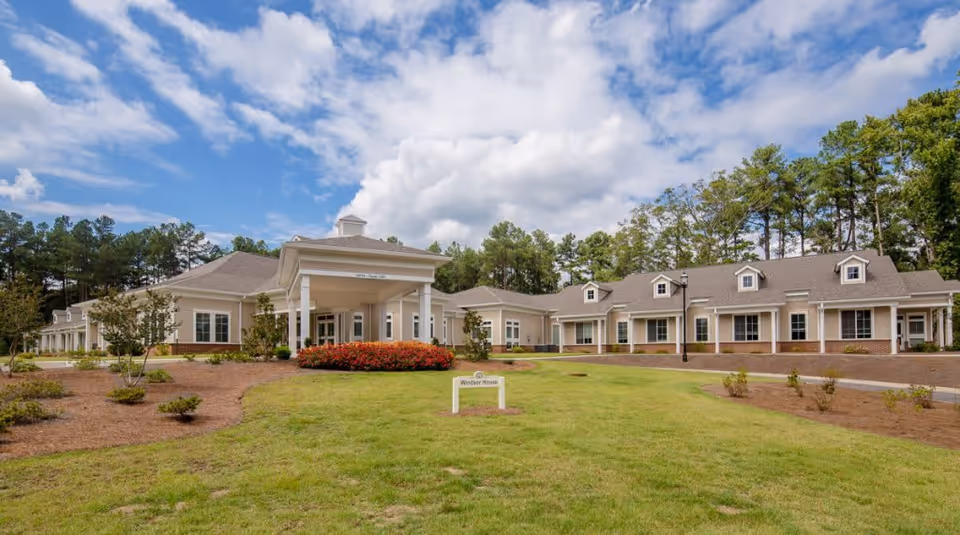 Exterior view of a single-story senior living facility building with a covered entrance, surrounded by a well-maintained lawn, small shrubs, and trees under a partly cloudy sky.