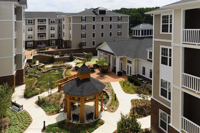 Courtyard of a senior living facility featuring a wooden gazebo, winding walkways, a pond, and surrounding multi-story buildings.