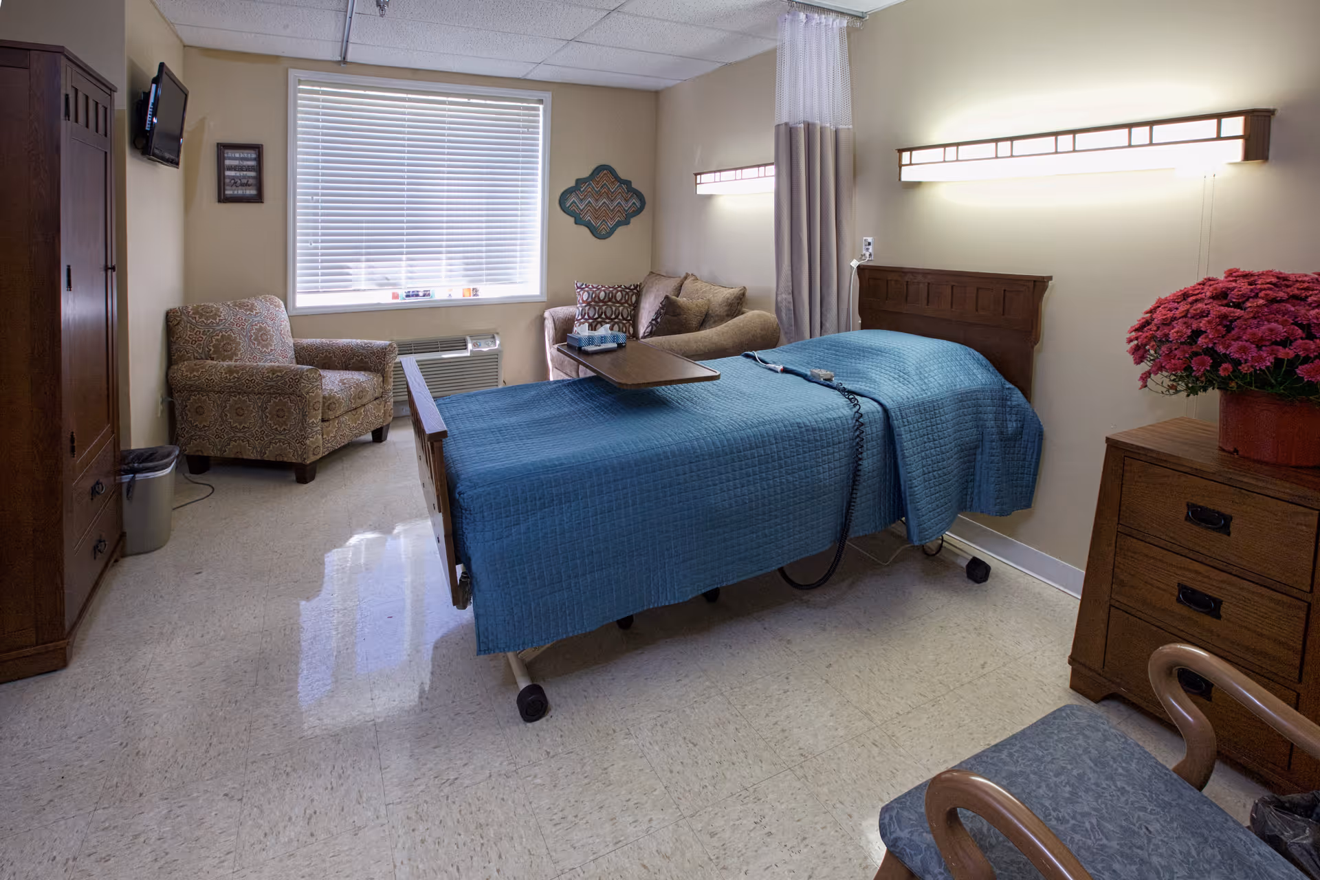A well-lit senior living facility bedroom with a hospital-style bed covered in a blue quilt, a wooden headboard, and a tray table. The room includes a patterned armchair, a small sofa with decorative pillows, a wooden dresser with a pot of pink flowers, a wall-mounted TV, and a window with blinds letting in natural light.