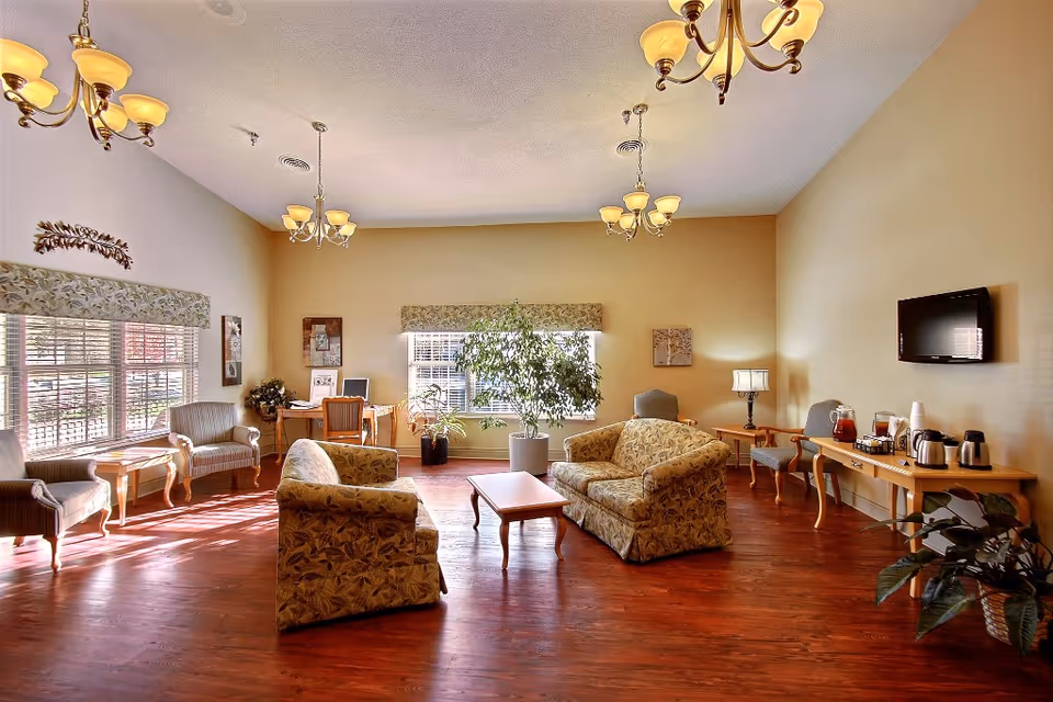Spacious sunlit lounge with patterned sofas and armchairs arranged around a coffee table, a wall-mounted TV, and a refreshment table.
