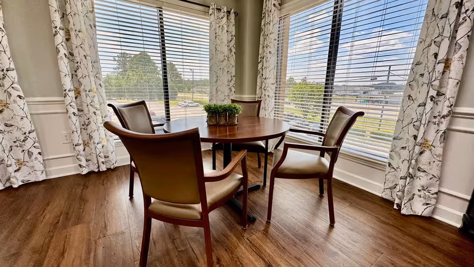 A small round wooden table with four cushioned chairs around it, placed near large windows with white floral curtains. Outside the windows, a parking lot and greenery are visible. The floor is wooden.