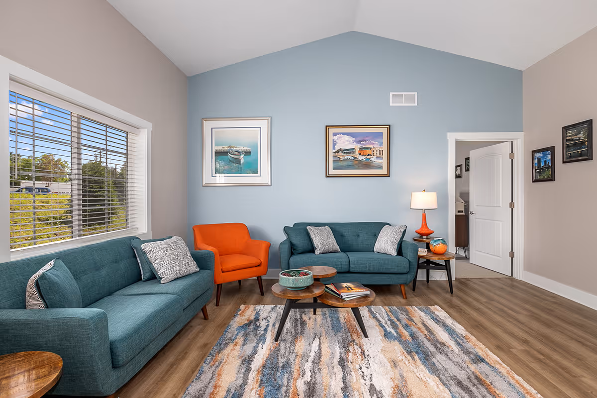 A cozy living room with two teal sofas and one orange armchair arranged around three nested wooden coffee tables on a multicolored rug. The walls are painted light blue and beige, decorated with framed paintings. A window with blinds lets in natural light, and a side table with an orange lamp is next to one sofa. The room has wood flooring and a white door leading to another room.