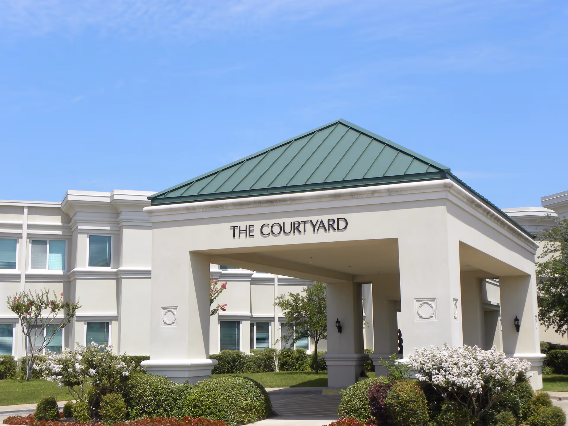 Exterior view of a building entrance with a green metal roof and a white archway labeled 'THE COURTYARD'. The surrounding area has neatly trimmed bushes, flowering plants, and trees under a clear blue sky.