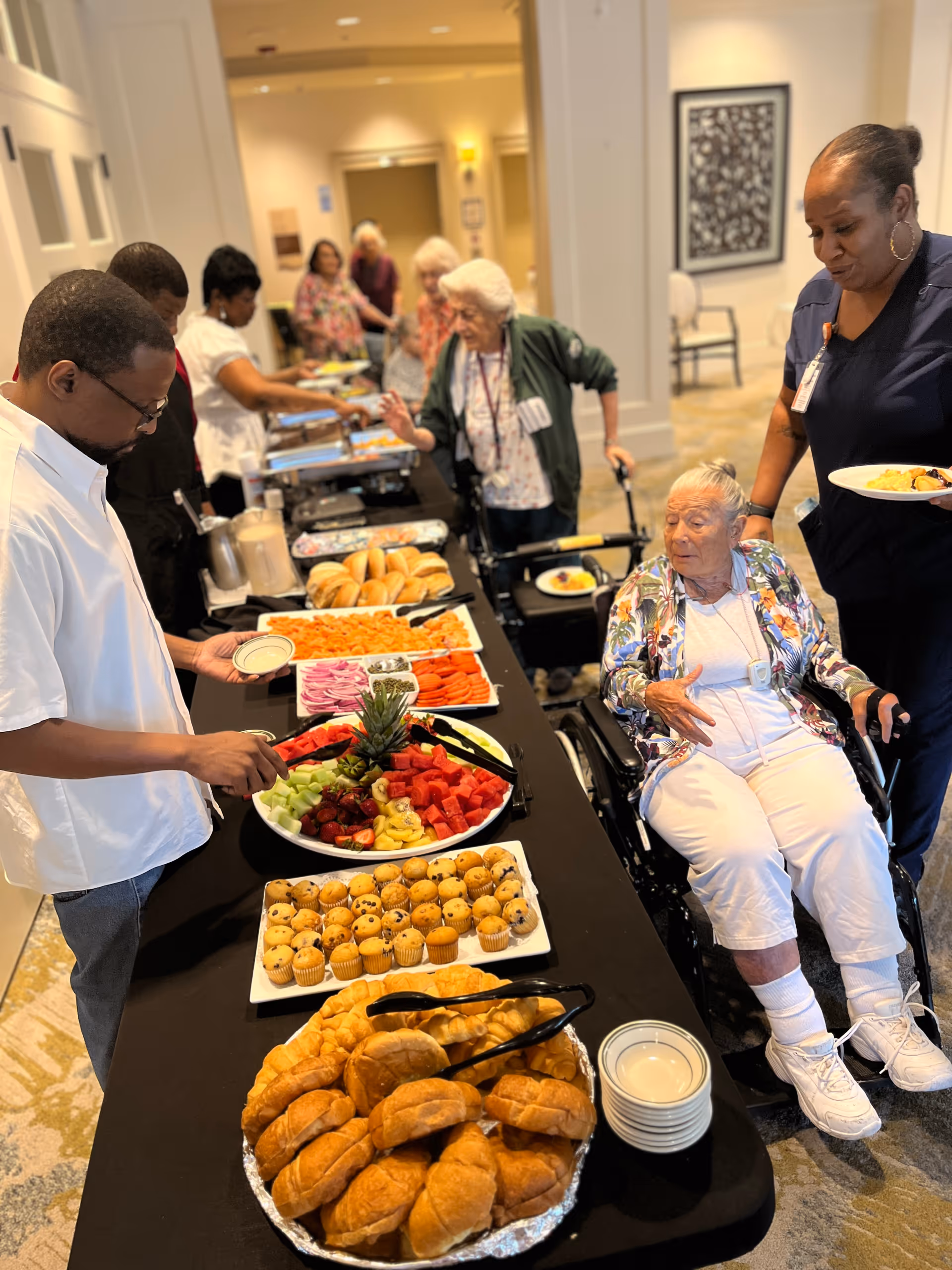 Residents and staff gather around a buffet table with rolls, muffins, fruit, and other dishes in a communal dining area.