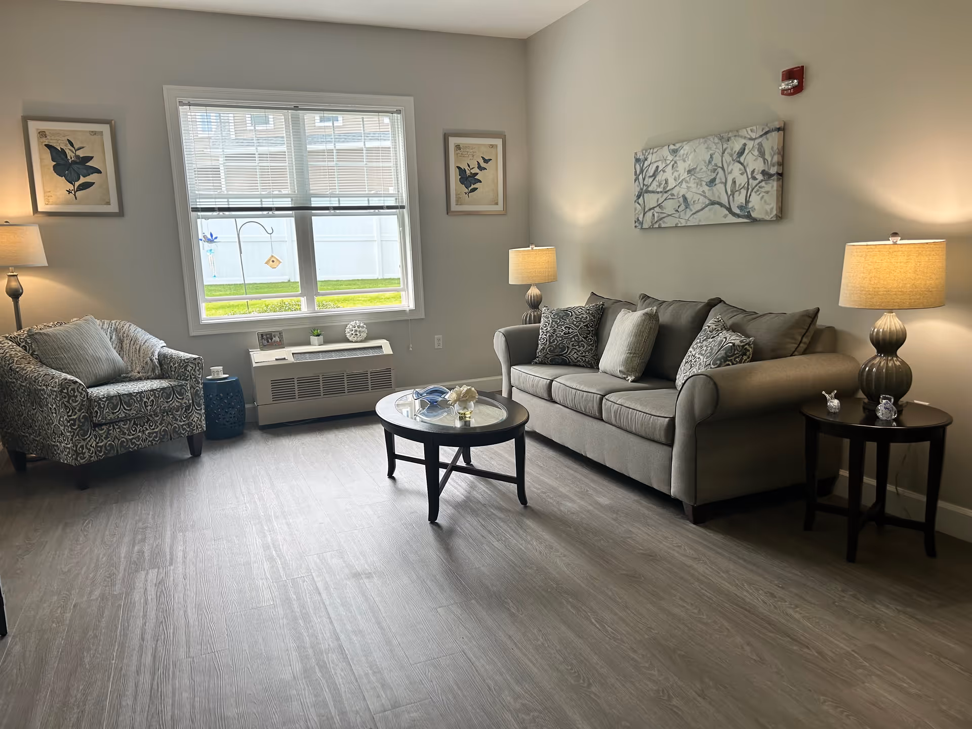 A cozy living room with a large window letting in natural light. The room features a patterned armchair with a pillow, a gray sofa with multiple cushions, two side tables each with a lamp, a round glass coffee table with decorative items, and framed artwork on the walls. The floor is wood-style laminate.