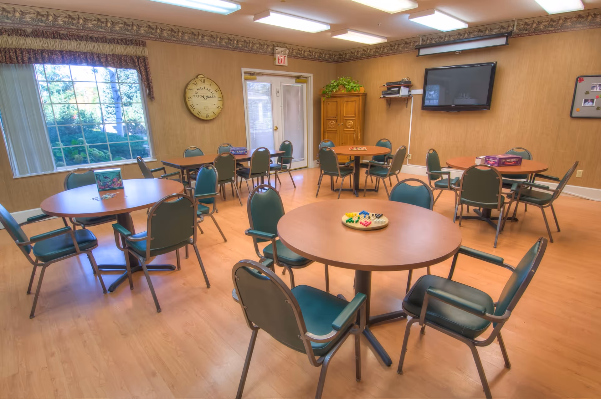Bright communal activity/dining room with several round tables and green chairs, a wall-mounted TV, and a large window.